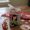 Pequeña caja de regalo blanca decorada con la imagen de la princesa Disney Mulán y un lazo rosa, usada para fiestas de cumpleaños o souvenirs.