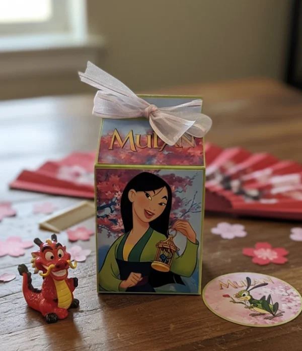 Pequeña caja de regalo blanca decorada con la imagen de la princesa Disney Mulán y un lazo rosa, usada para fiestas de cumpleaños o souvenirs.